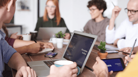 Close-up of a group of professionals using laptops, tablets, and smartphones in a conference room during a meeting, showcasing a BYOD (Bring Your Own Device) environment.