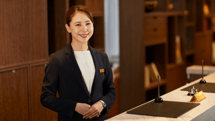 A hotel receptionist in a navy blazer stands smiling at the front desk, ready to greet guests, with a service bell and pen stand on the marble counter in a warmly lit lobby.