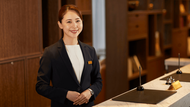 A hotel receptionist in a navy blazer stands smiling at the front desk, ready to greet guests, with a service bell and pen stand on the marble counter in a warmly lit lobby.
