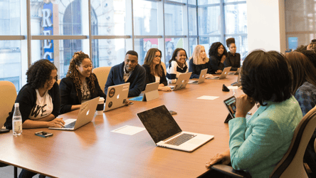 A diverse group of professionals seated around a large wooden conference table in a modern office. They are engaged in discussion, working on laptops and tablets, with large windows offering a city view in the background. The atmosphere reflects teamwork and collaboration in a hybrid work environment.