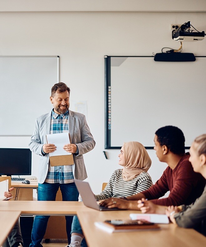 A college teacher talking to a group of students during a class in the classroom. With Boom technology, classroom interactions are elevated, fostering effective communication and engaging teaching sessions.